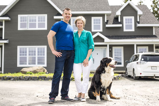 A man and woman standing in front of their house with a dog by their side.